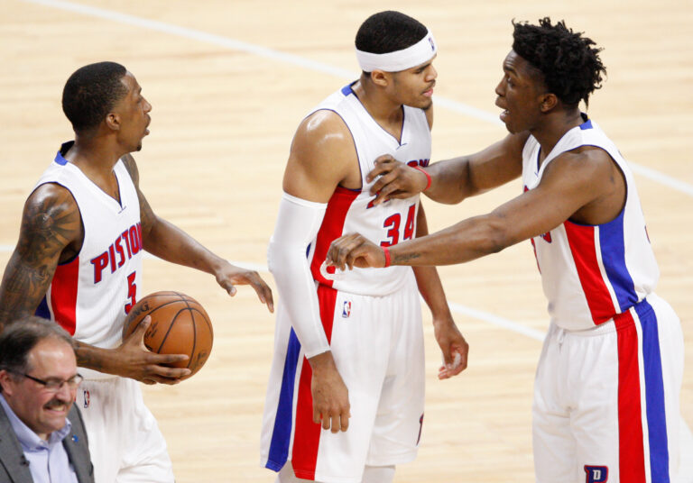Three Detroit Pistons players in white uniforms celebrate on the court, arms linked and smiling.