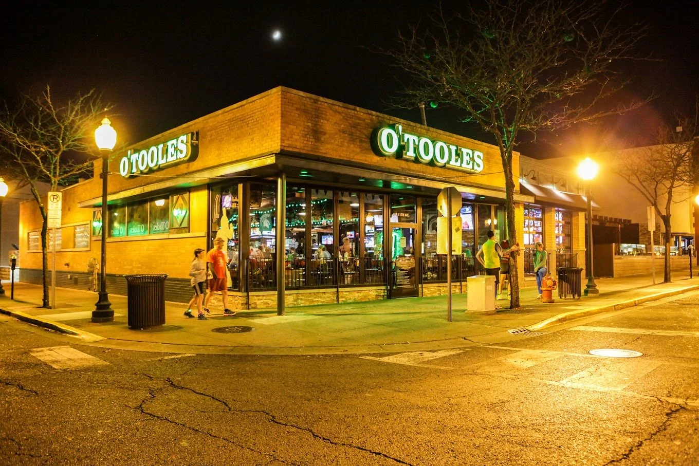 Nighttime street corner exterior of O'Toole's pub with glowing green sign, brick façade, and warm interior lights visible through large windows. Several pedestrians on the sidewalk.
