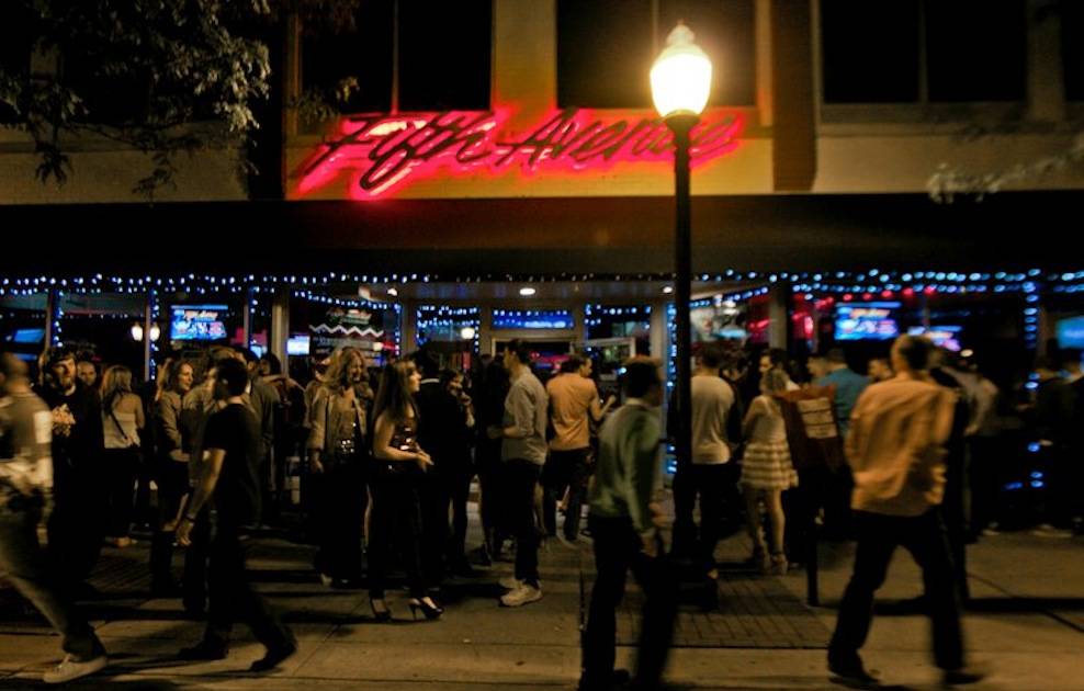 Night scene outside a nightlife venue with a pink neon sign, a streetlamp, and a group of people mingling under blue string lights.