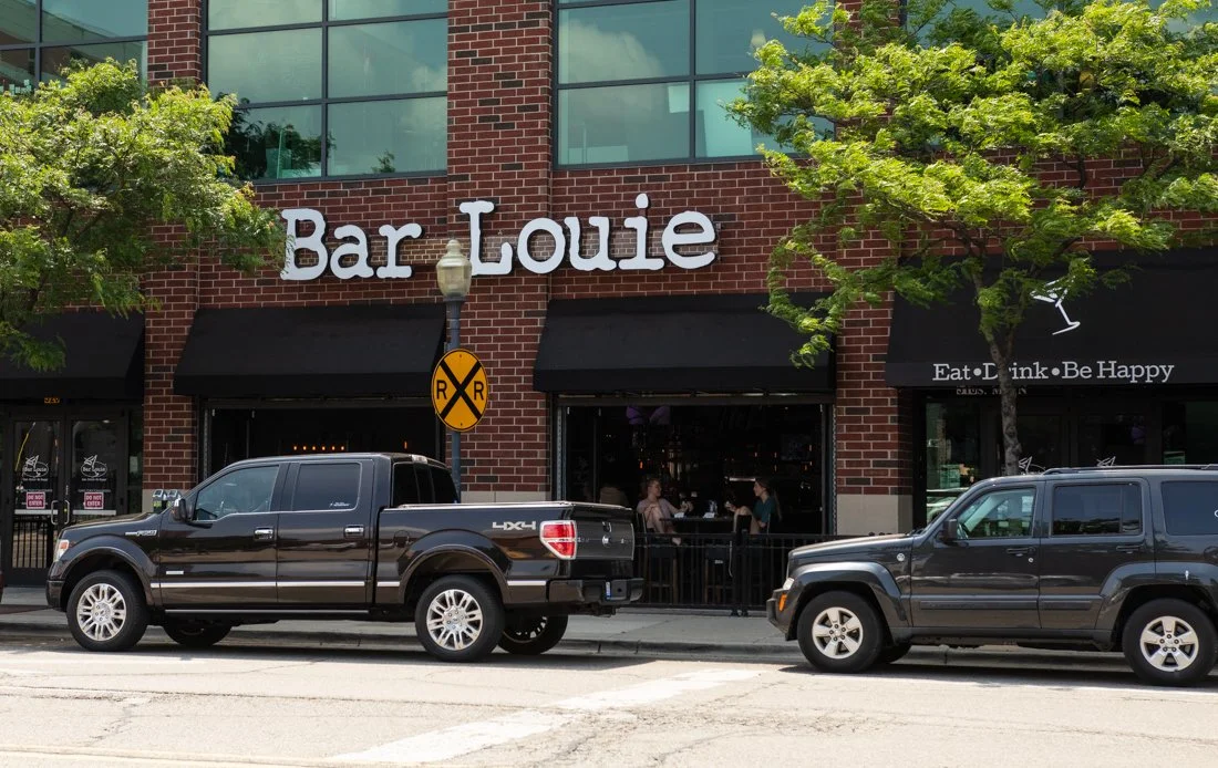 Exterior view of Bar Louie restaurant on a brick building, with two trucks parked along the street and pedestrians inside.
