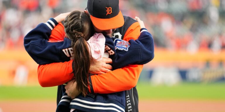 A man in a Detroit Tigers cap and orange jacket hugs a girl in a navy jacket on a baseball field, stands blurred in the background.