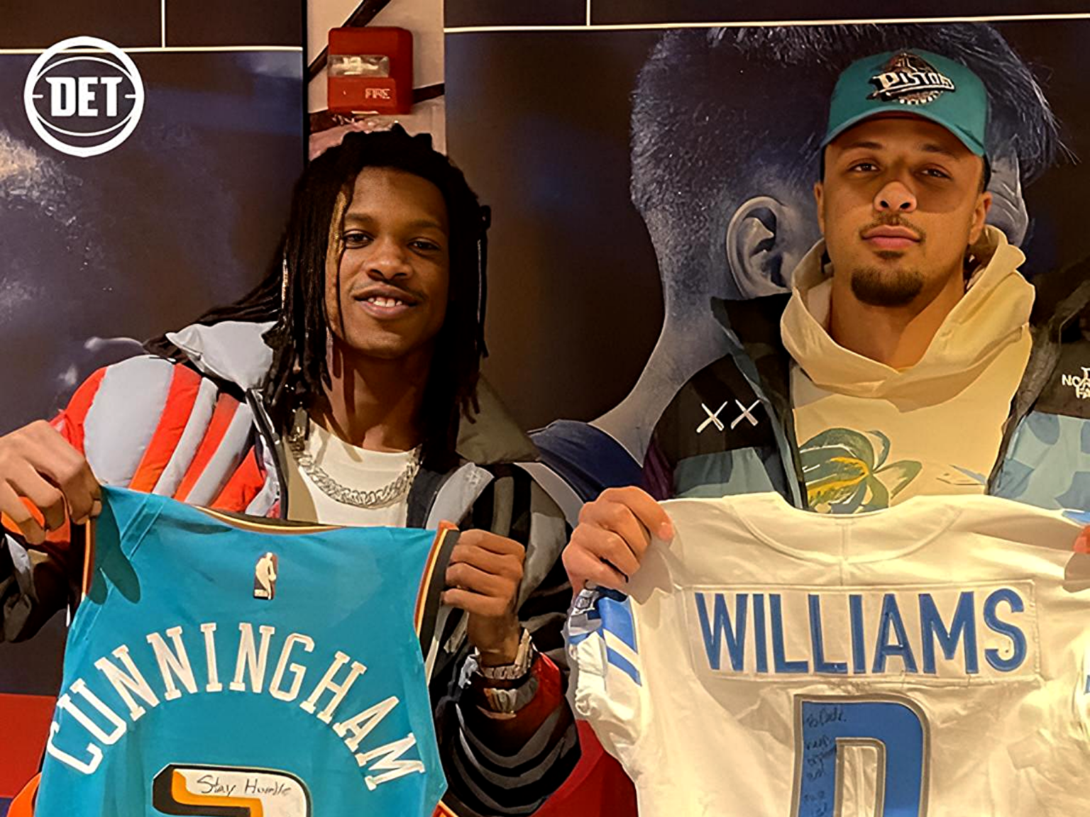 Two young men stand together holding large banners with bold names, one reading GUNNINGHAM and the other WILLIAMS, in front of a dark display backdrop.