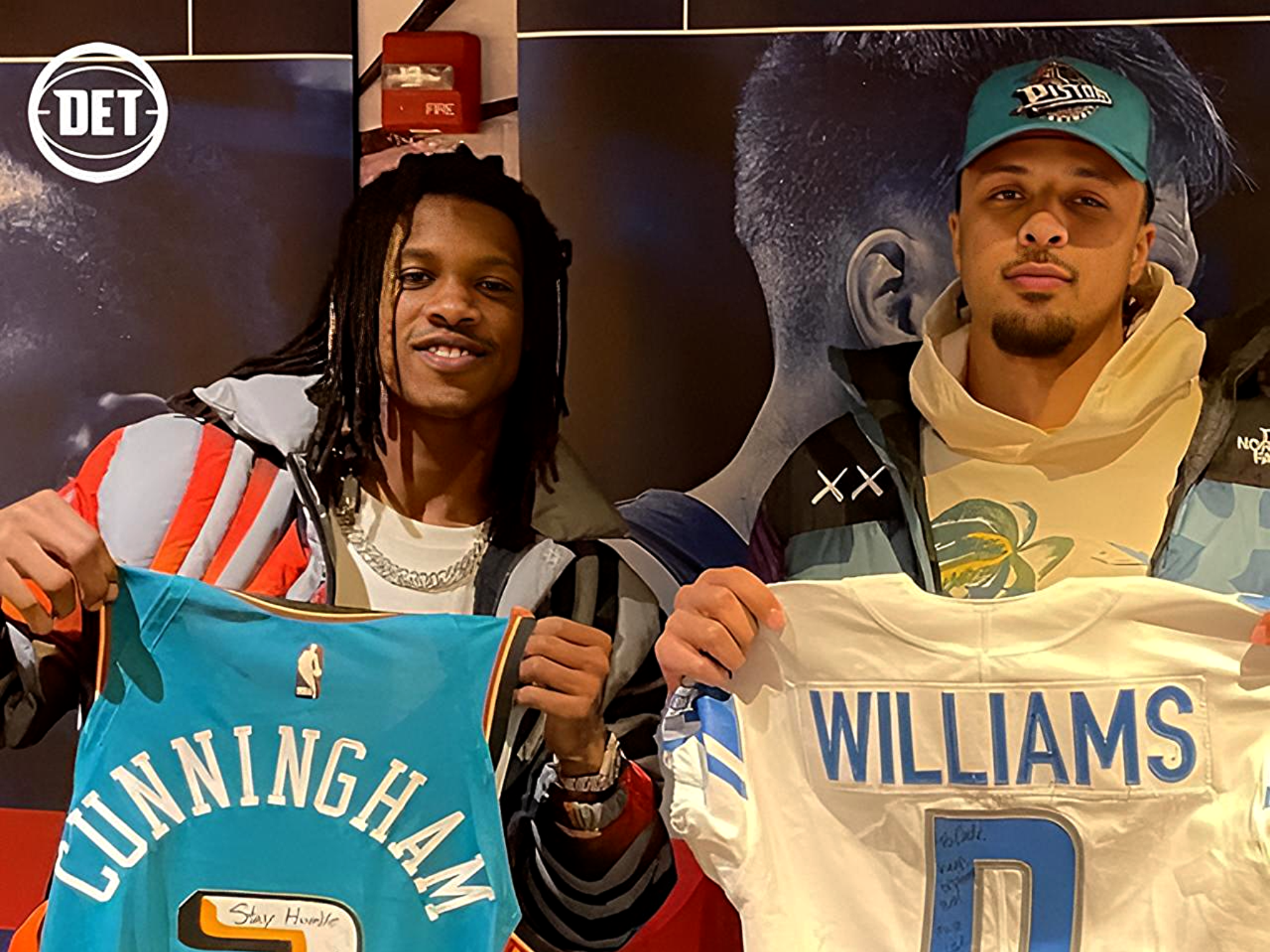 Two young men stand together holding large banners with bold names, one reading GUNNINGHAM and the other WILLIAMS, in front of a dark display backdrop.