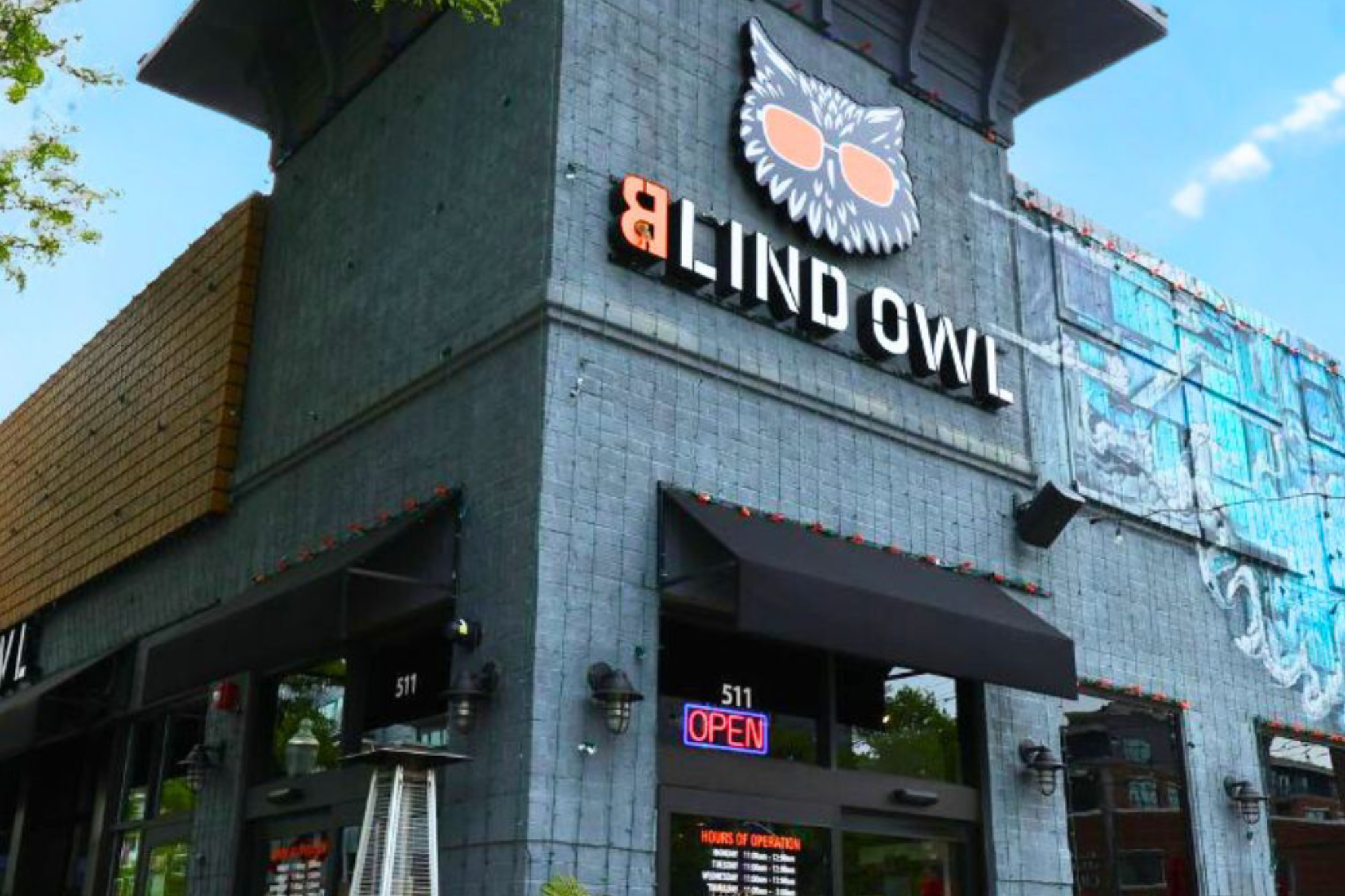 Front of a gray brick storefront with a large owl logo sign and an illuminated OPEN sign.